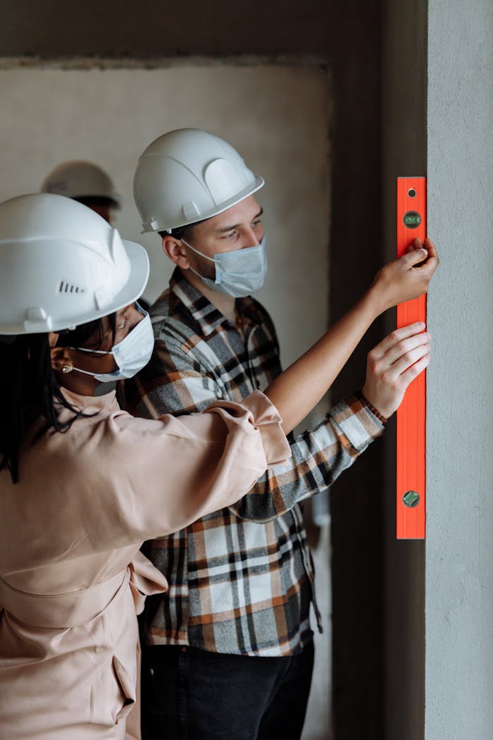 Two engineers measuring wall with a level indoors, wearing face masks and helmets for safety.